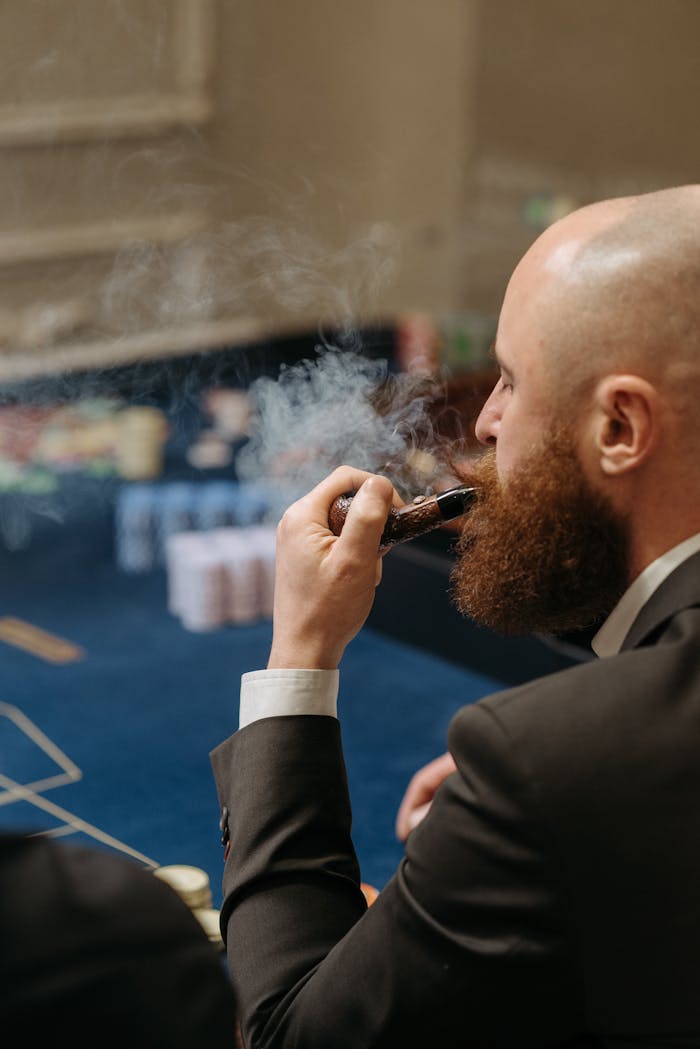 Bald man with beard smokes a pipe inside a casino, surrounded by a sophisticated atmosphere.
